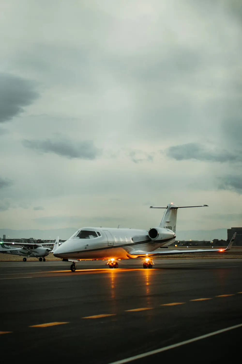 A business jet with its landing lights on, waiting on a wet ramp under a heavy evening sky