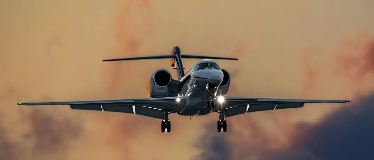 A business jet on approach, landing lights on, against a sunset sky